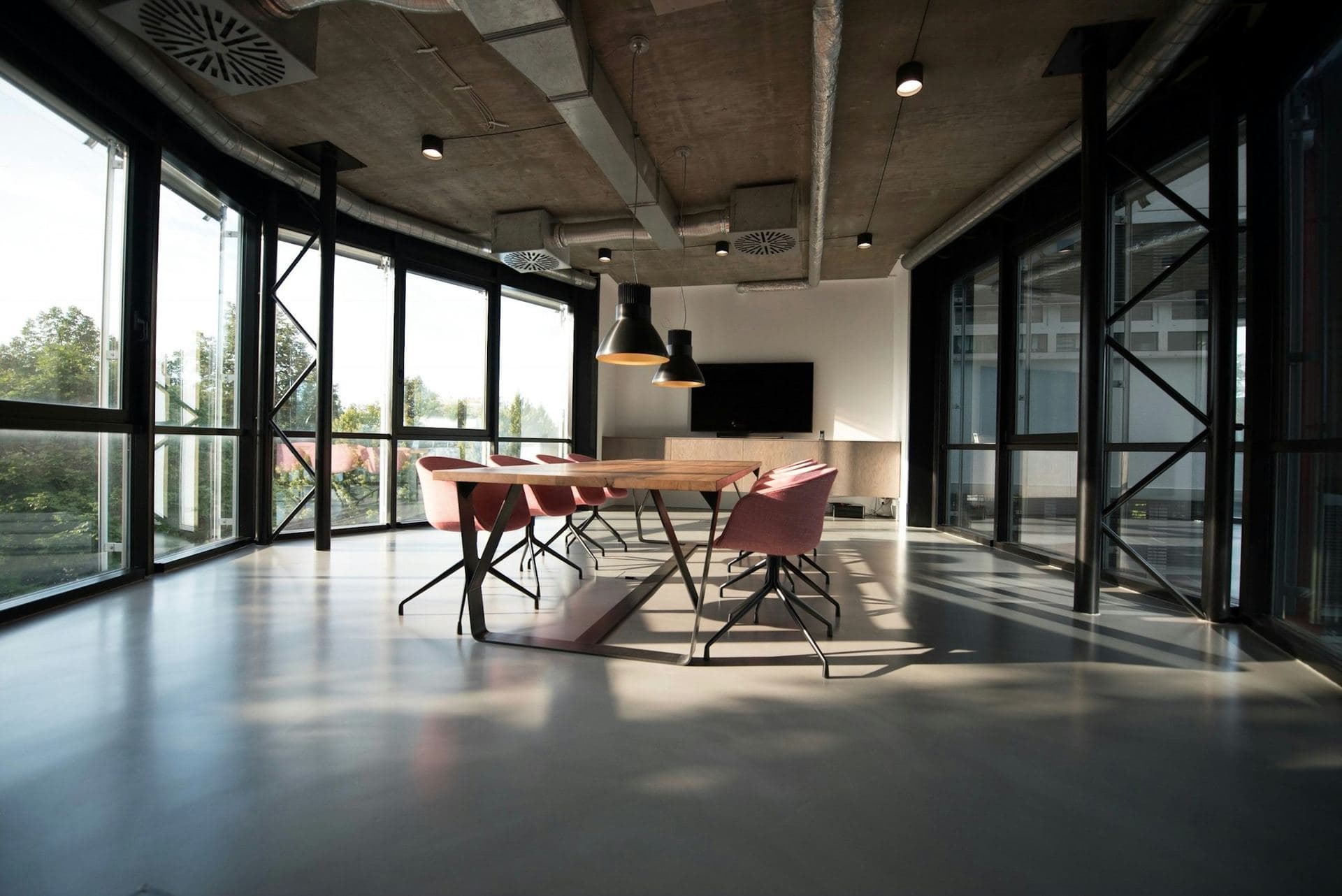 Espace de travail vide aux grandes baies vitrées, longue table en bois sous des suspensions, lumière naturelle au sol