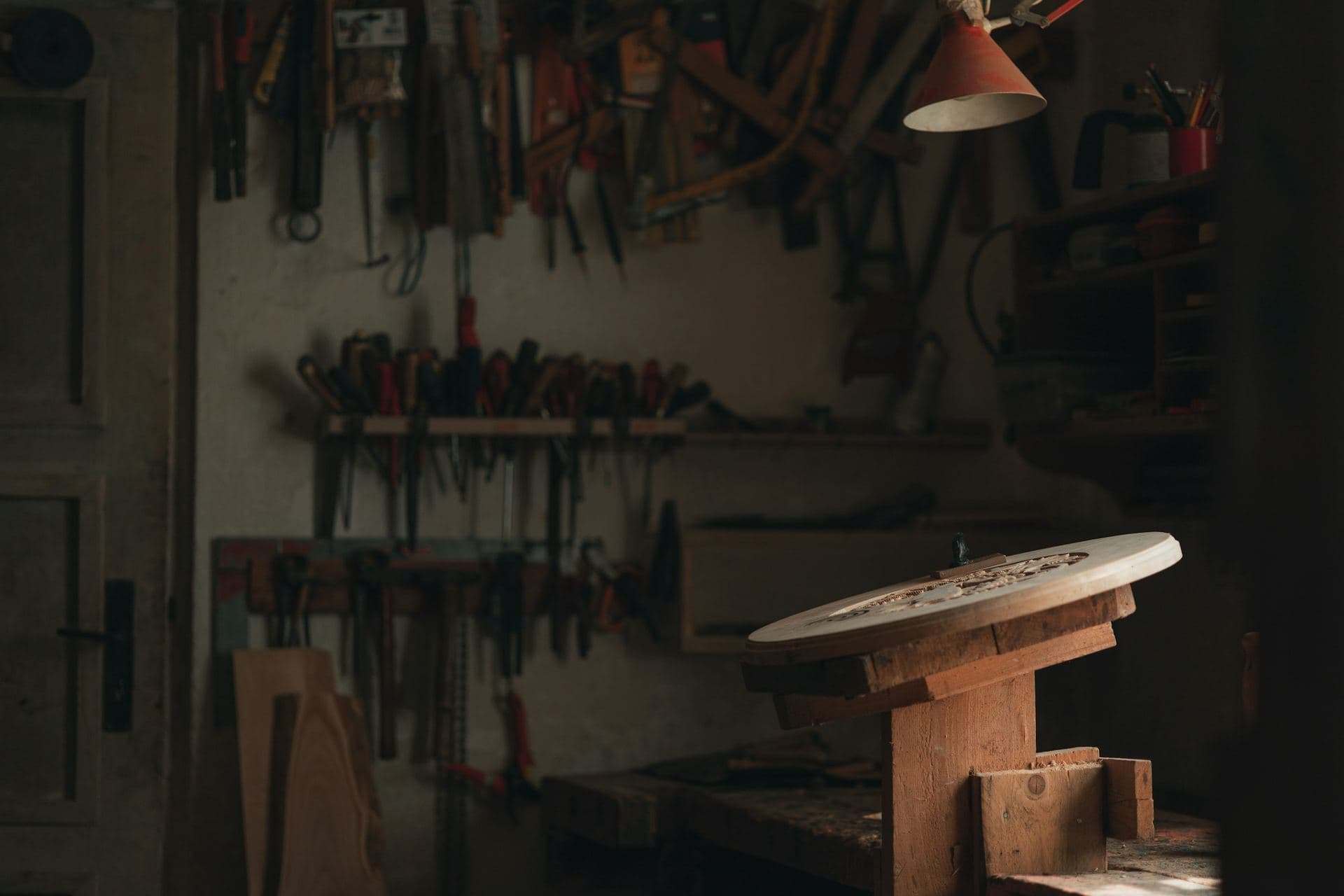 Atelier d'artisan en fin de journée, établi et outils au mur sous la lumière chaude d'une lampe, porte à gauche