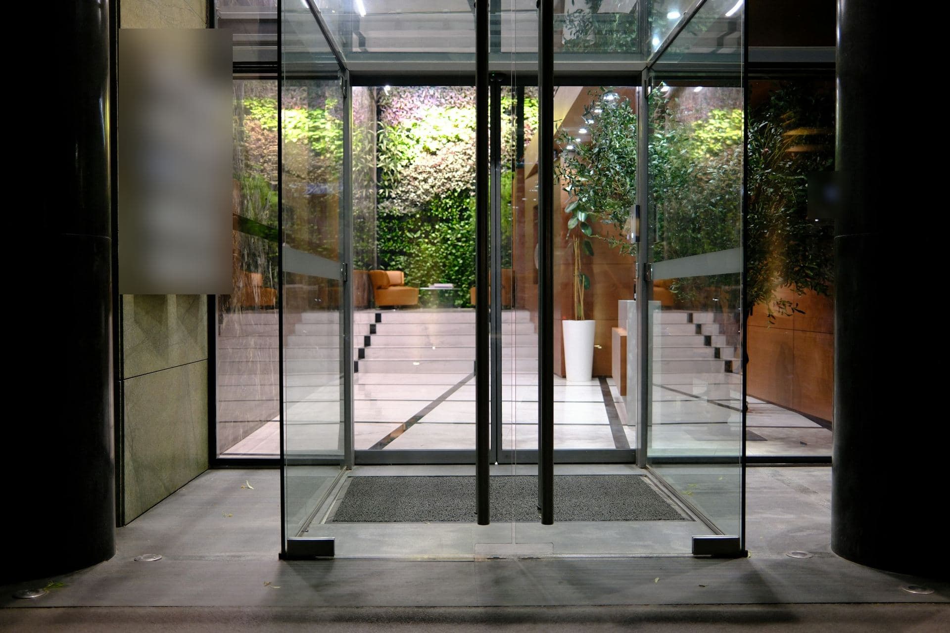 Portes vitrées ouvertes sur le hall éclairé d'un immeuble de bureaux, marches de marbre et mur végétal au fond
