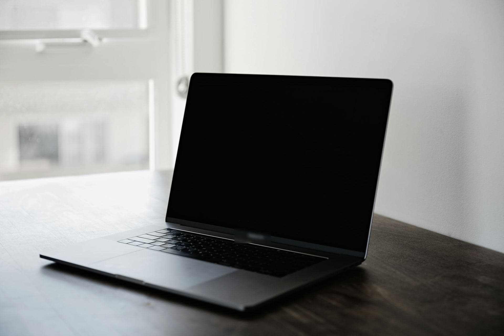 Ordinateur portable ouvert, écran éteint, vu de trois quarts sur une table sombre près d'une fenêtre
