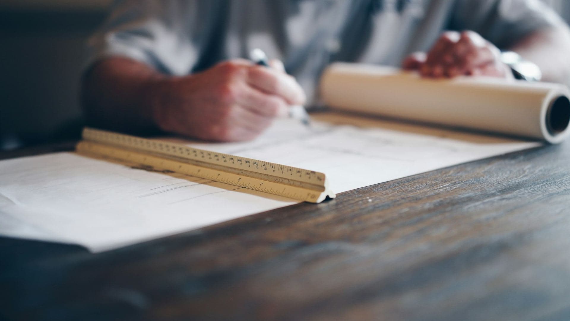 Mains traçant un plan d'architecte avec une règle d'échelle posée sur la table, rouleau de calque en arrière-plan