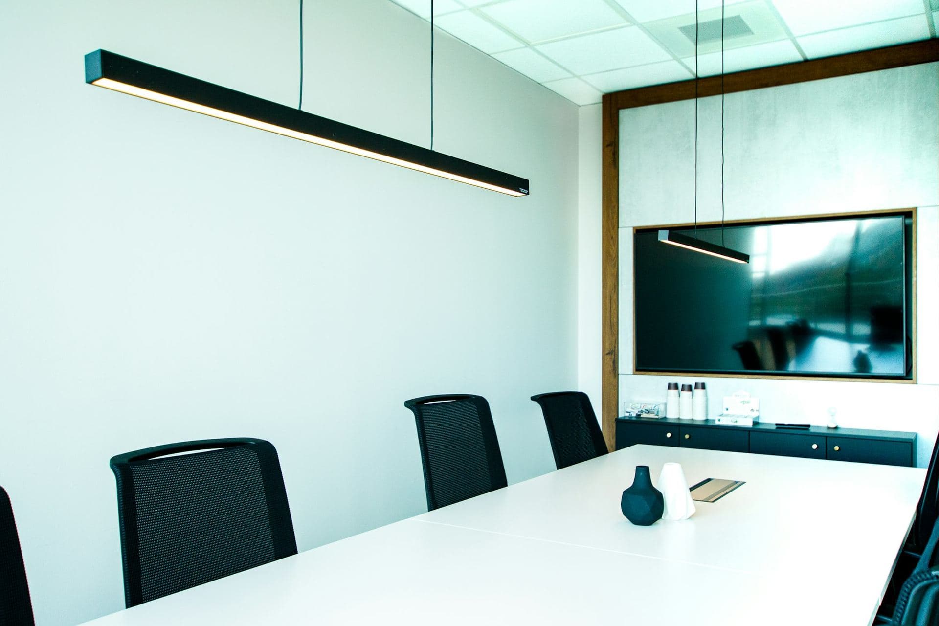 Salle de réunion vide, longue table blanche, fauteuils noirs et luminaire linéaire suspendu